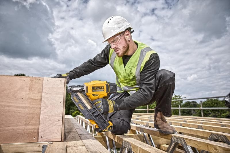 Person uses a DEWALT nail gun DCN930N on wooden boards at an outdoor construction site, wearing safety gear.