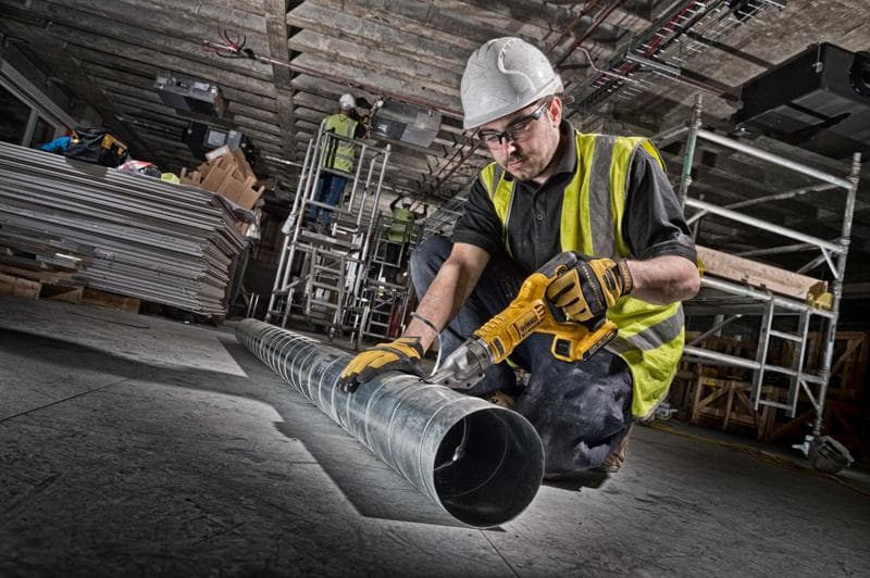 Worker uses DEWALT cordless metal shear on duct pipe at construction site. Safety gear and tools visible.