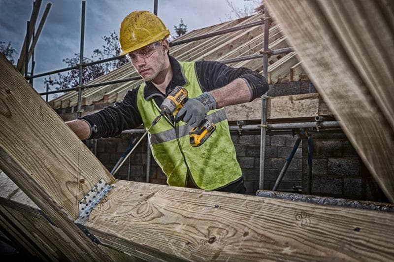 Person using a DEWALT cordless drill on wooden beams at a construction site, wearing safety gear and high-visibility vest.