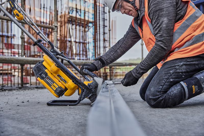 Worker uses yellow DEWALT tool to saw metal at construction site, wearing safety gear and orange vest.